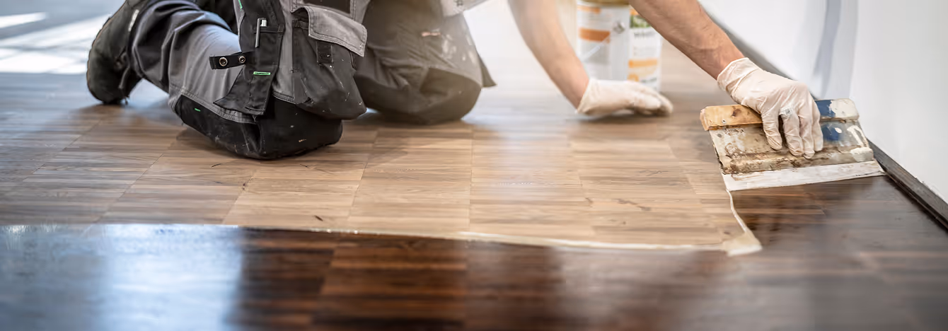Person kniet auf einem Boden und trägt eine Beschichtung mit einem Spachtel auf einem Holzfußboden auf.