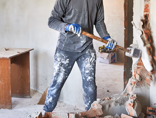 Handwerker mit blauer Handschuhen und baufarbverschmierten Hosen schlägt mit einem Vorschlaghammer eine Wand ein.