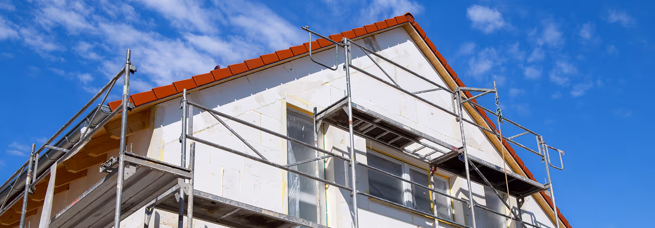 Oberer Teil eines Hauses mit weißer Fassade und rotem Dach, umgeben von Gerüstbau unter blauem Himmel mit wenigen Wolken.
