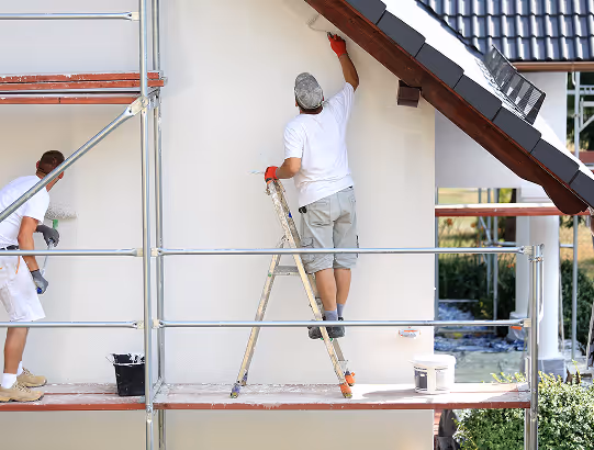 Zwei Handwerker renovieren die Außenwand eines Hauses mit Gerüst, einer streicht die Wand, der andere arbeitet links.