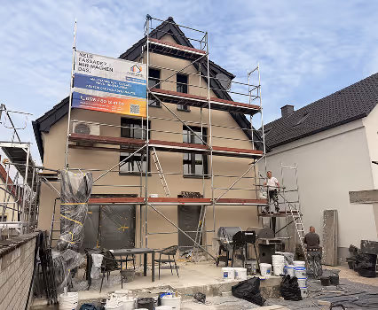 Haus mit Gerüst und Bauarbeitern beim Fassadenanstrich an einem sonnigen Tag.