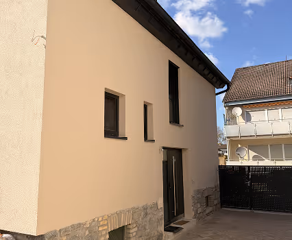 Haus mit hellgelber Fassade, kleinen Fenstern und einem dunklen Eingang unter blauem Himmel mit einigen Wolken.
