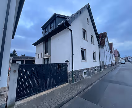 Wohnhaus mit weißer Fassade und schwarzem Tor an einer gepflasterten Straße unter bewölktem Himmel.
