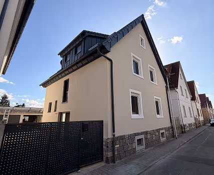 Zweigeschossiges modernes Wohnhaus mit hellbeiger Fassade, Dachgaube und schwarzem Metallzaun an einer ruhigen Straße.