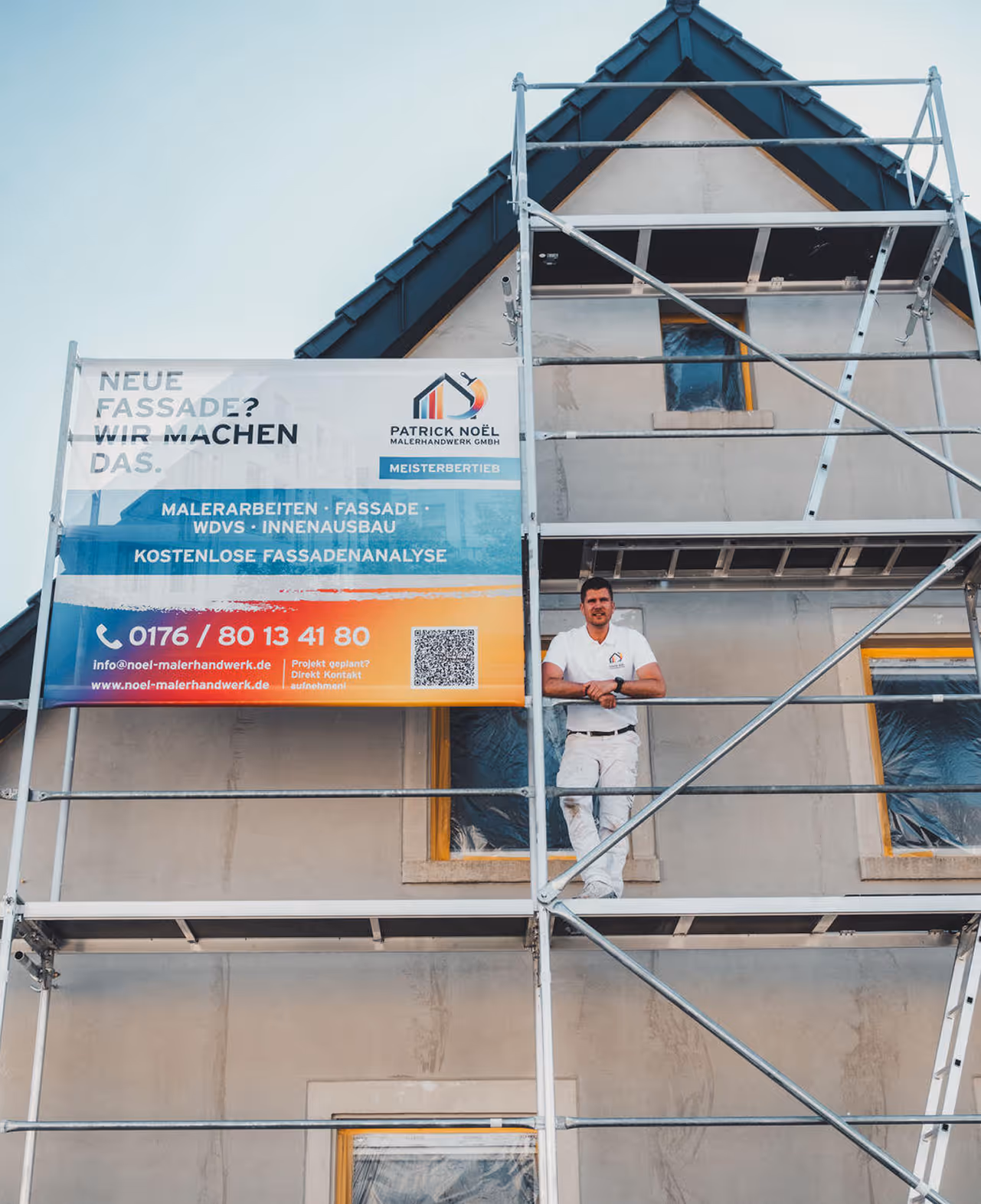 Maler auf Gerüst vor einem Haus mit Werbebanner für Fassadenarbeiten von Patrick Noël Malerhandwerk GmbH.