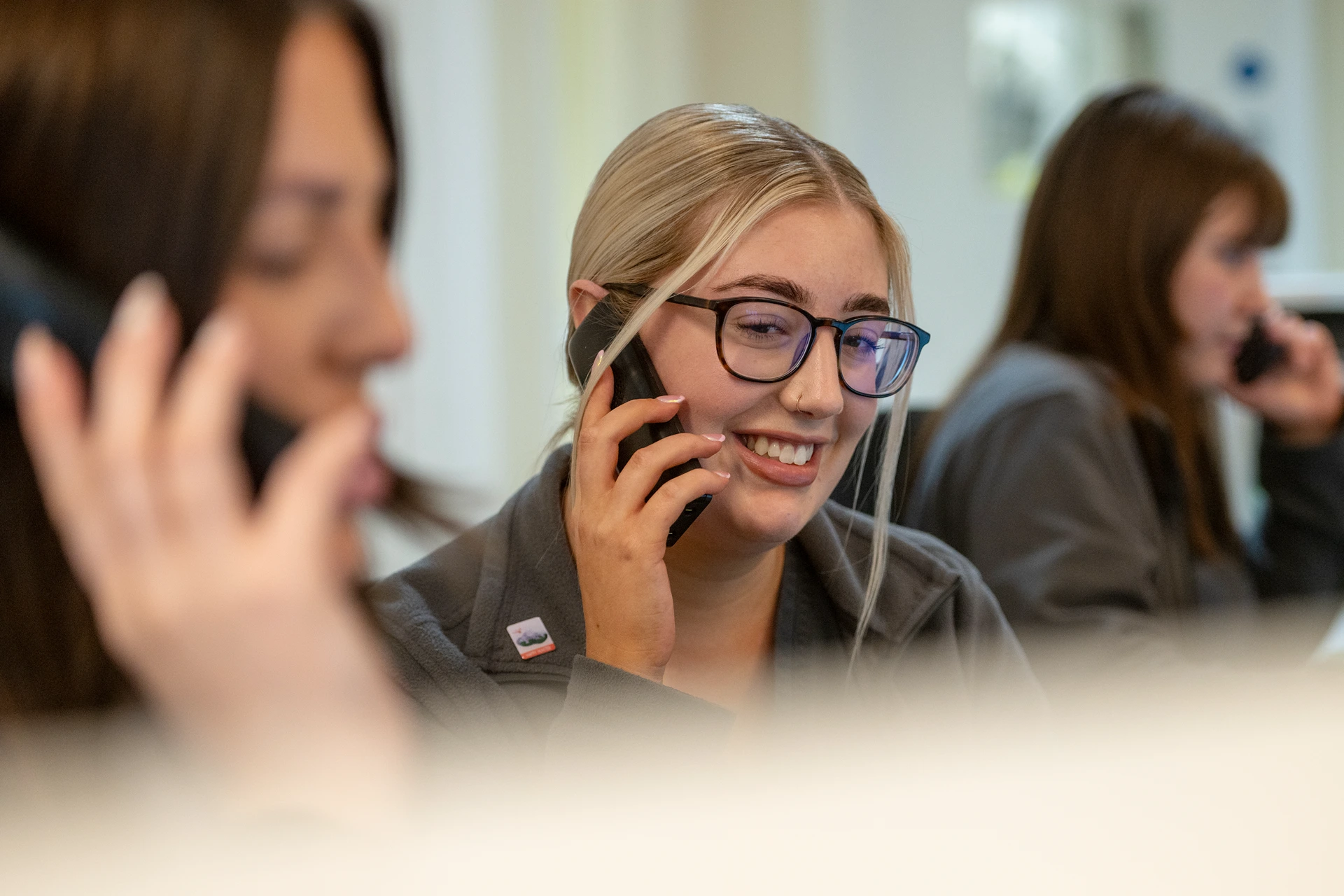 Smiling woman with glasses talking on a phone in an office with two blurred colleagues also on phones.