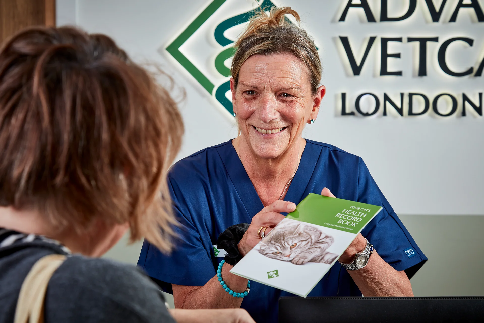 Veterinary professional in blue scrubs smiling and showing a cat health record book to a customer.