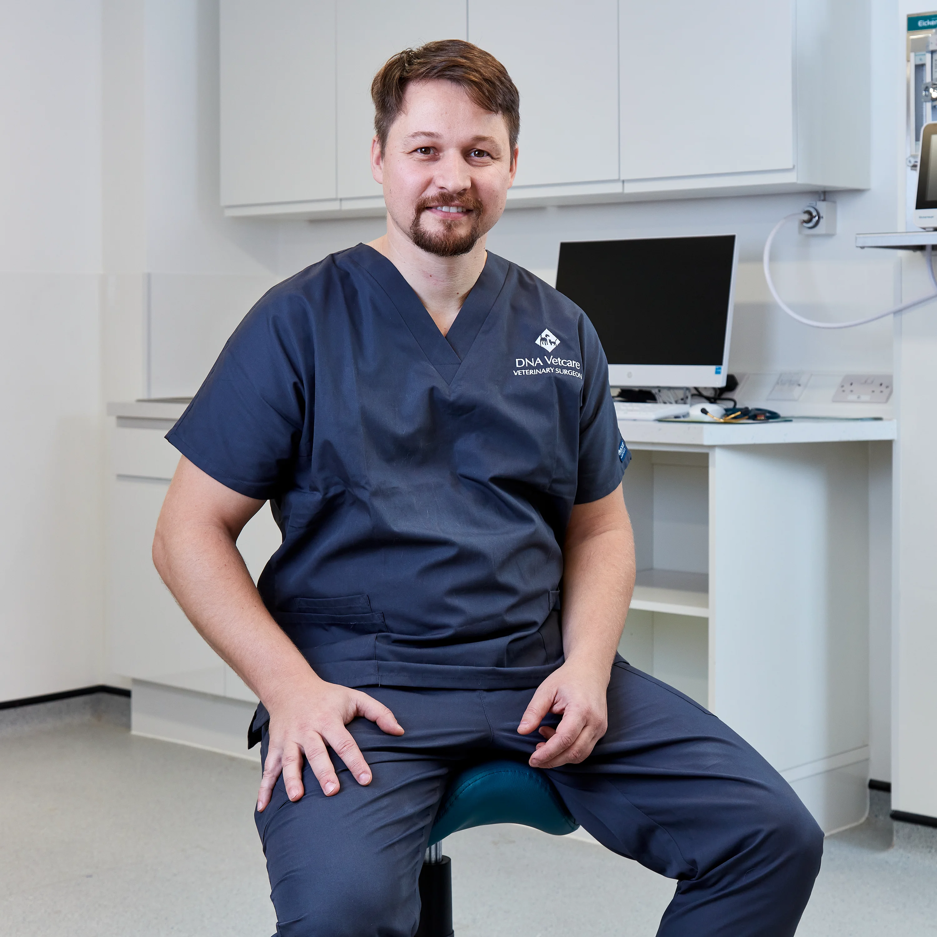 Male veterinarian in navy scrubs sitting on a stool in a veterinary clinic with a computer in the background.