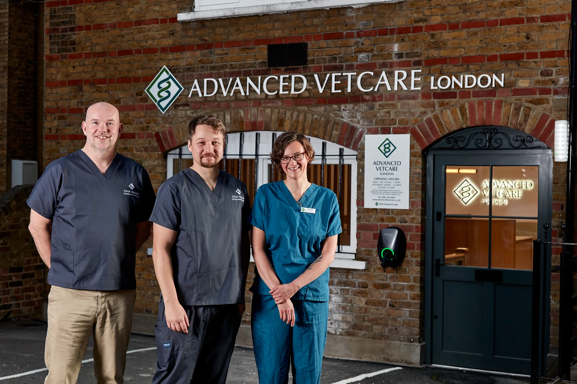 Three veterinary staff in scrubs standing in front of the Advanced Vetcare London clinic entrance at night.
