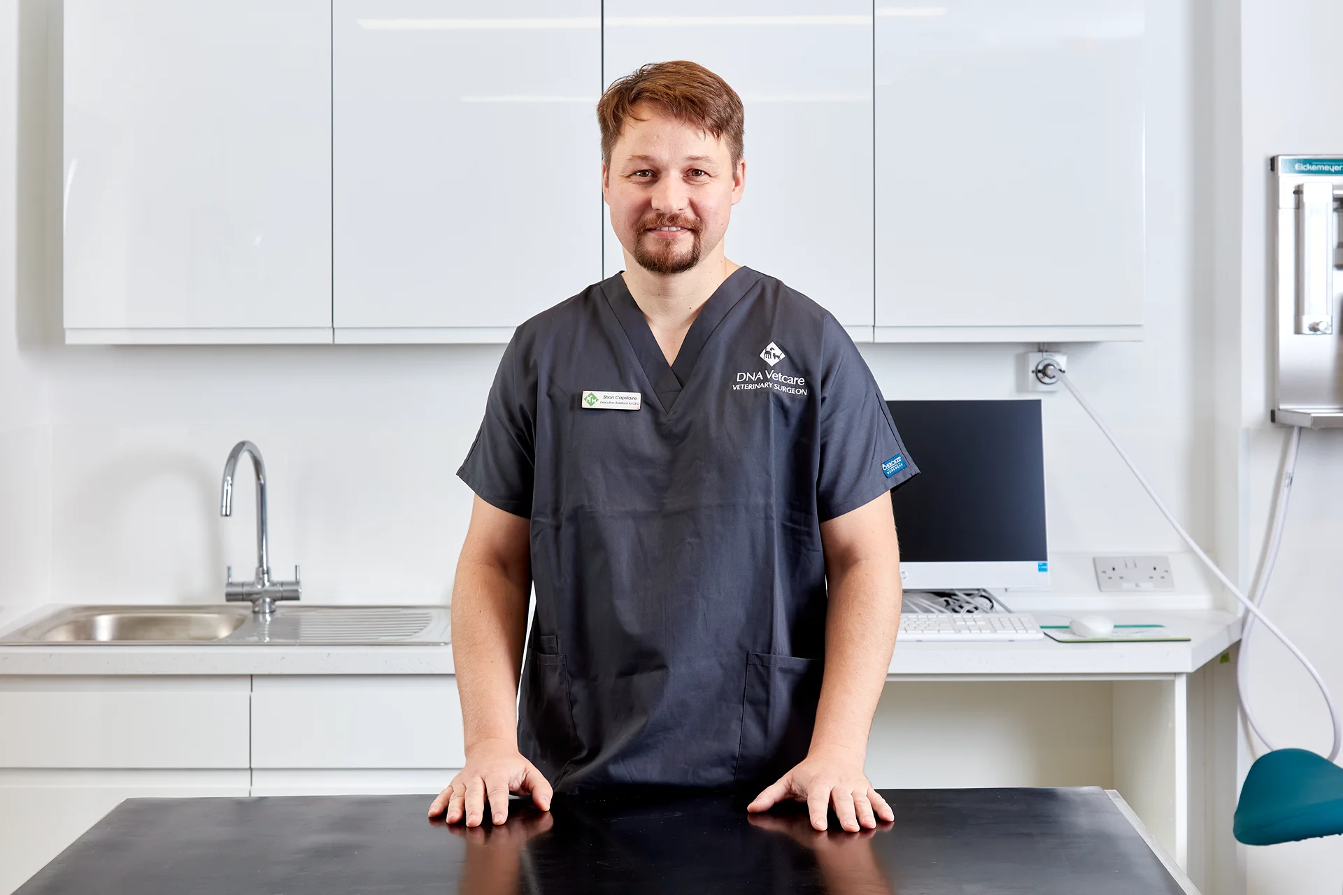 Male veterinary surgeon in dark scrubs standing behind an exam table in a clinical room.