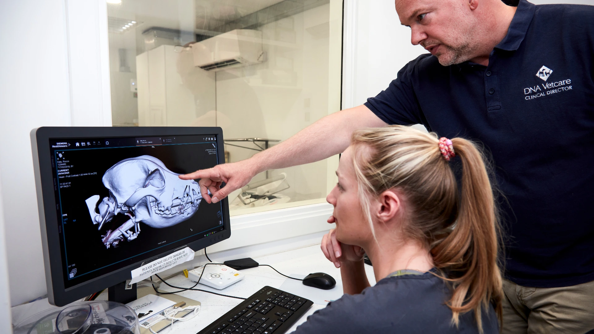 Veterinary professional pointing at a dog skull X-ray on a computer screen while explaining to a seated colleague.