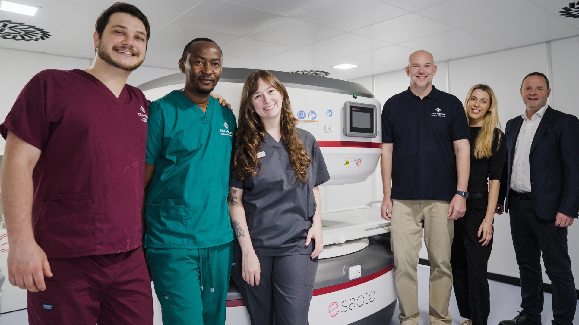 Six medical professionals standing and smiling in front of an Esaote MRI machine in a clinical setting.