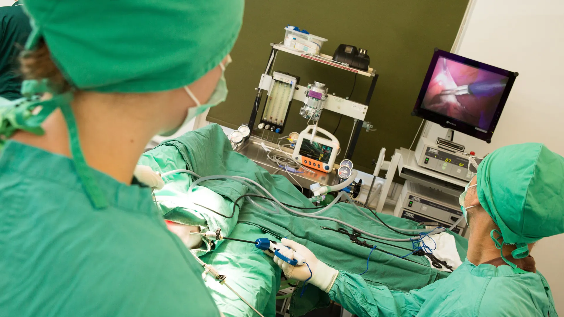 Surgeons in green scrubs performing laparoscopic surgery while watching a monitor displaying the internal view.