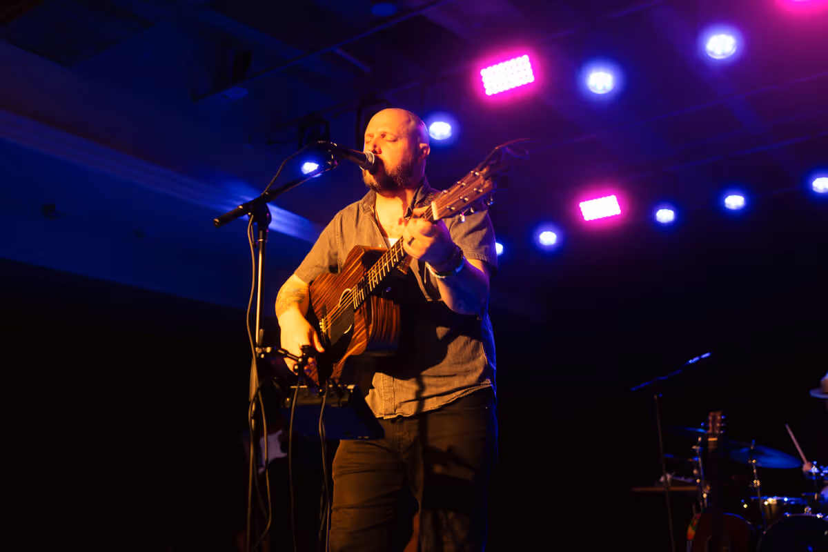 Male musician singing and playing an acoustic guitar on stage under purple and blue lights.