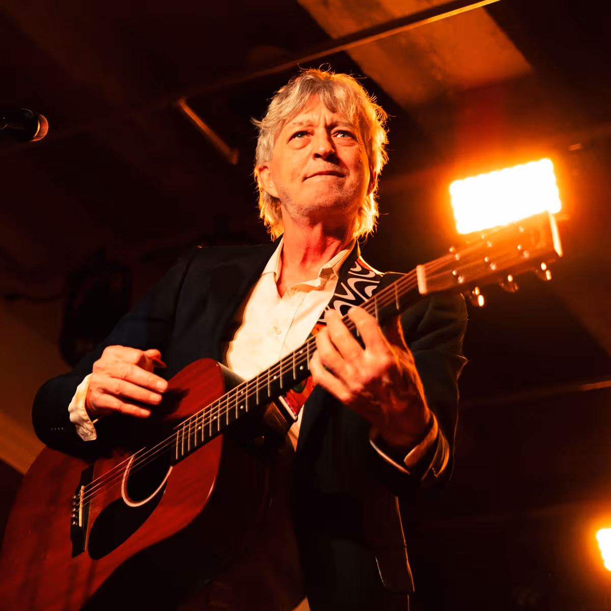 Older man playing a red acoustic guitar under warm stage lighting.