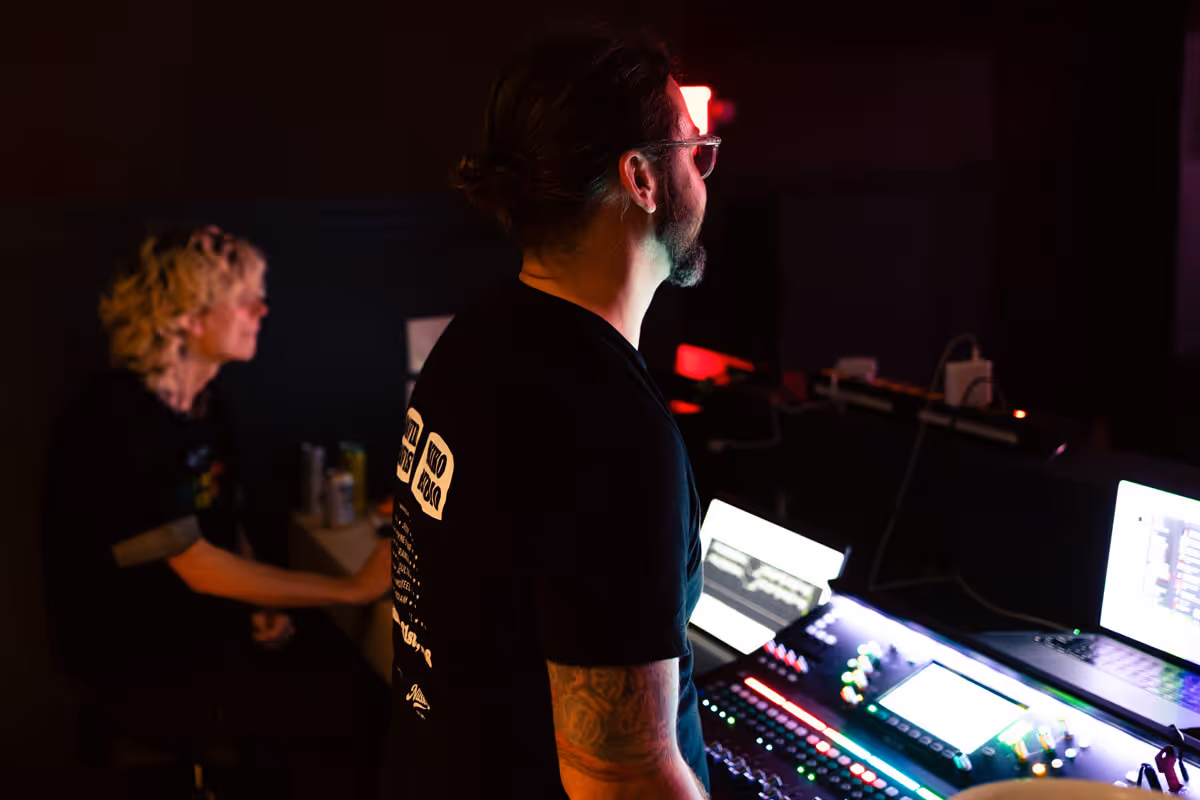 Two people in a dimly lit room operating a sound mixing console with illuminated controls.