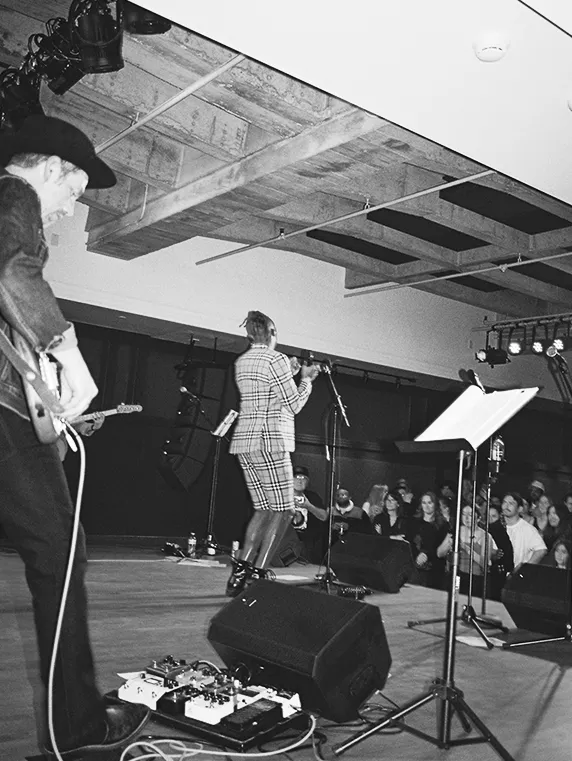 Black and white photo of a live music performance with a singer in plaid outfit and guitarist on stage, and an audience watching.