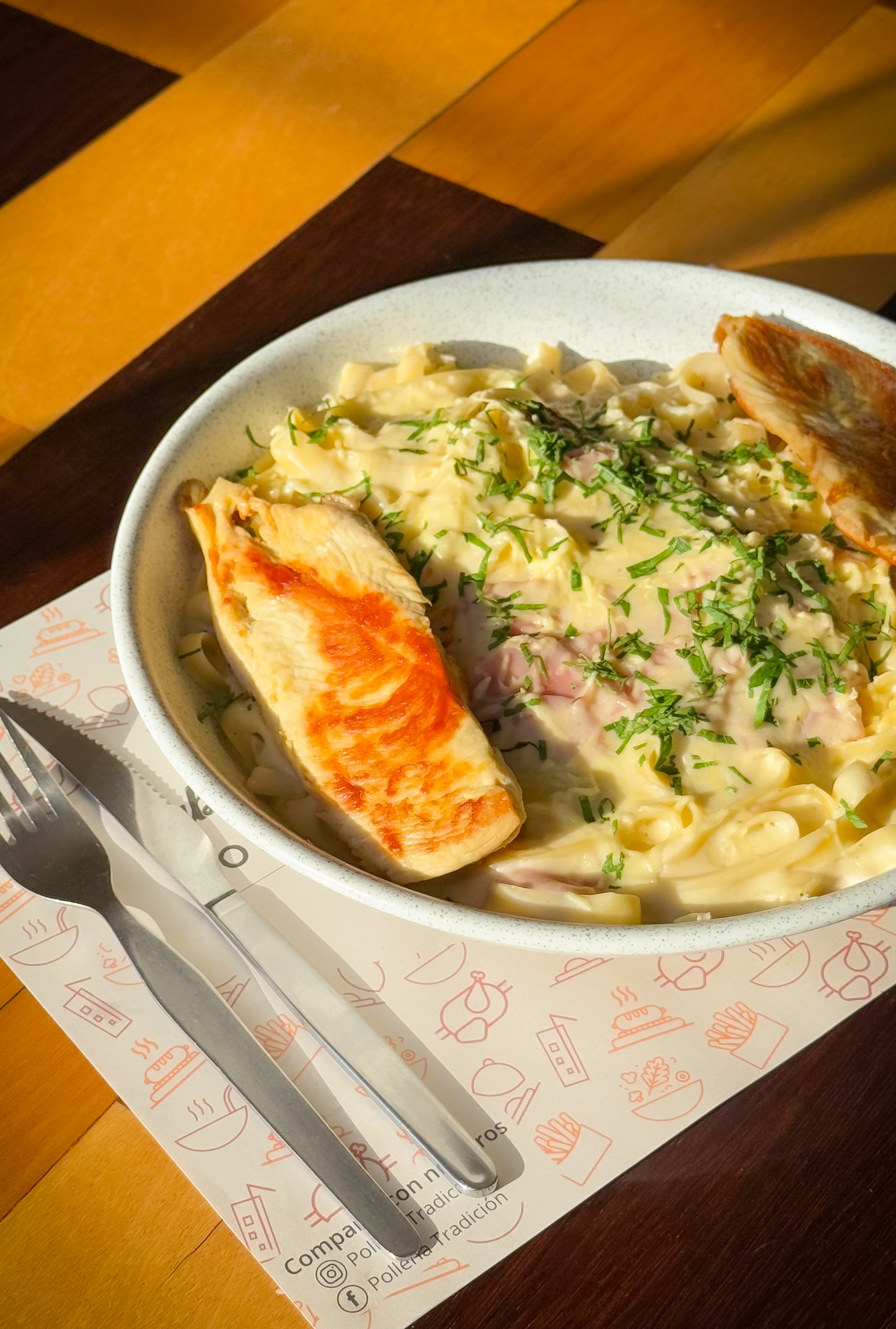 Un tazón de pasta cremosa con hierbas frescas, servido con dos piezas de pechuga de pollo a la parrilla. Junto al tazón se colocan un tenedor y un cuchillo.