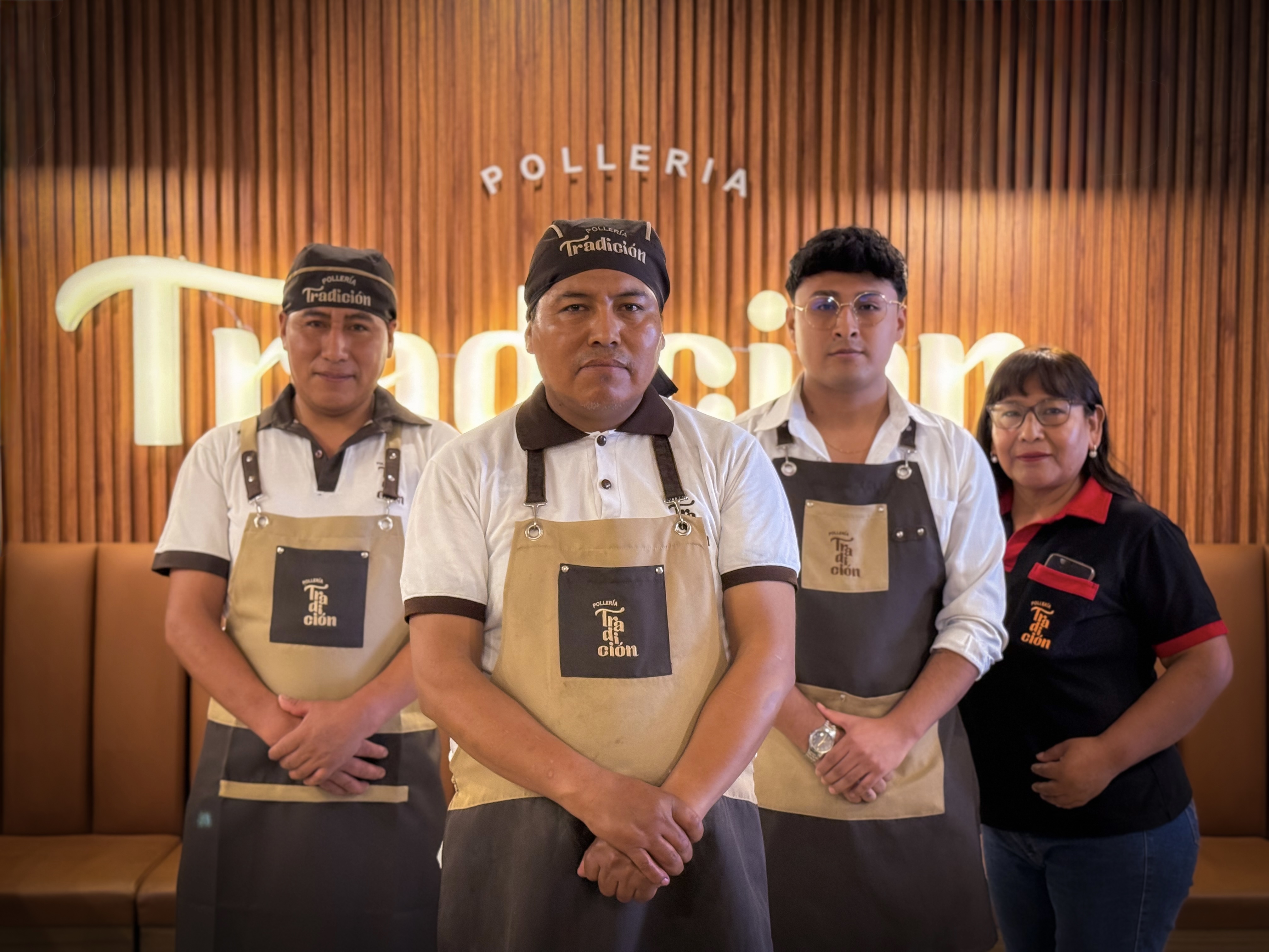 Cuatro miembros del personal del restaurante Tradición, tres con delantales y sombreros beige a juego y uno con una camisa negra y roja, están de pie frente a la cámara frente a una pared revestida de madera con el logotipo del restaurante Tradición.