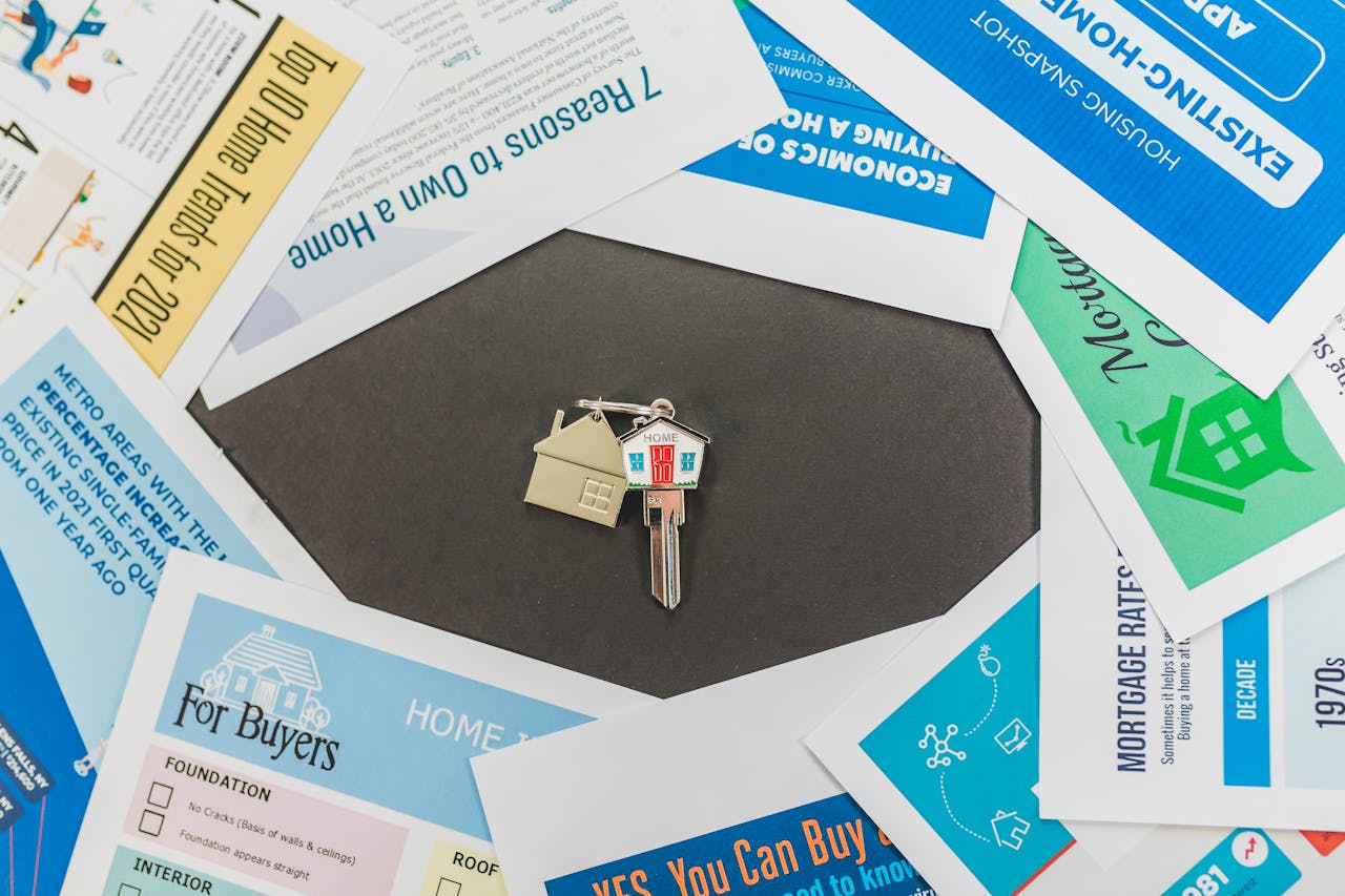 House-shaped keychain attached to a key placed on a black surface, surrounded by scattered home-buying and real estate documents and guides.