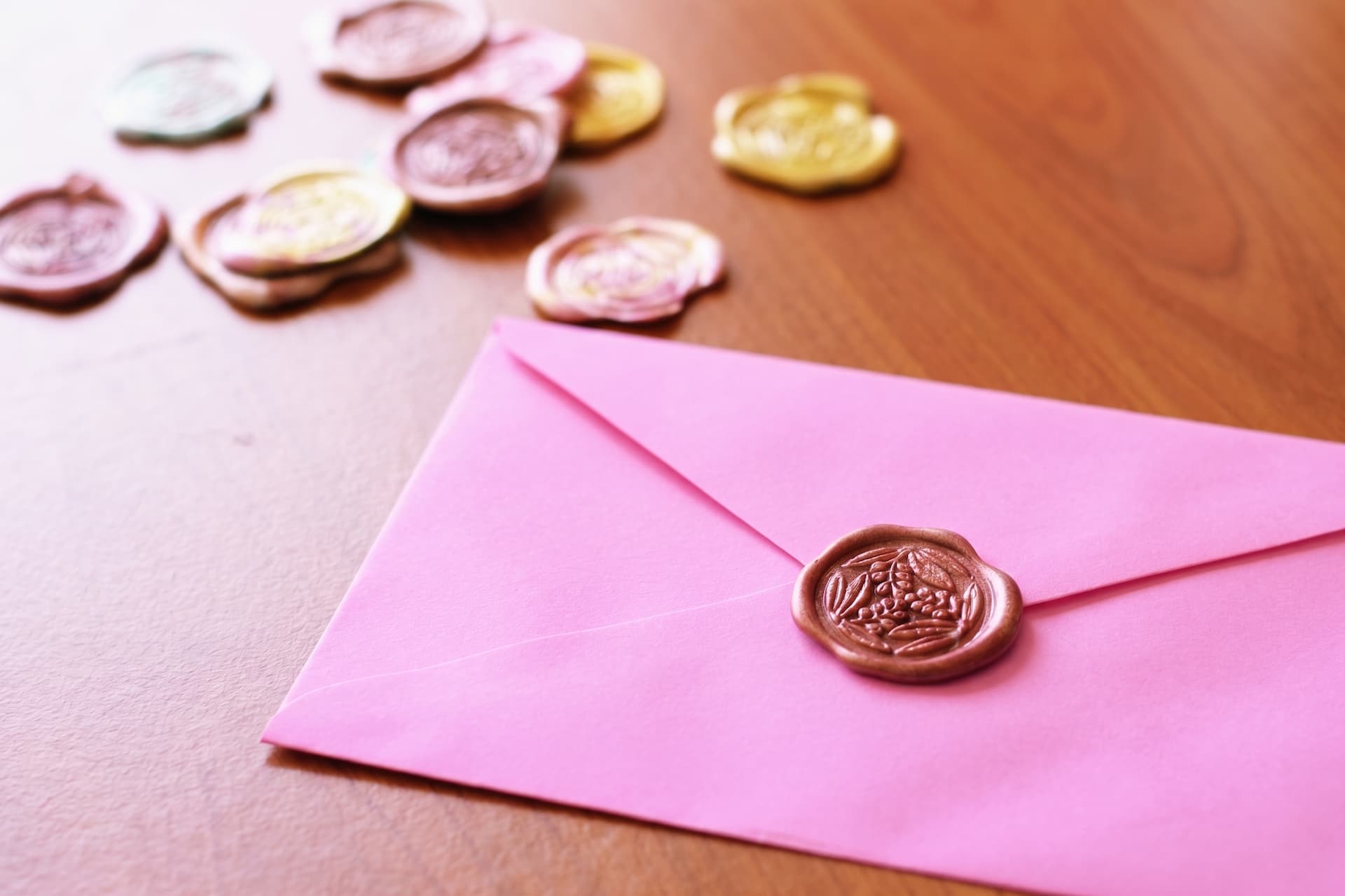 Pink envelope with a rose-gold floral wax seal on a wooden desk, with scattered wax seals behind it.