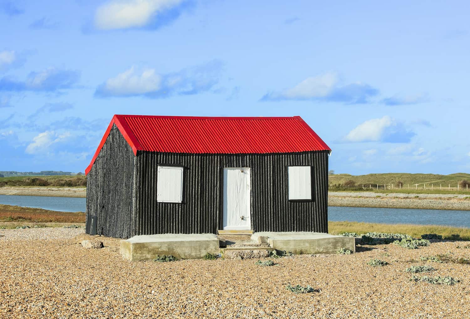 Non-standard construction coastal building with corrugated black siding, red metal roof, and white doors beside a waterway