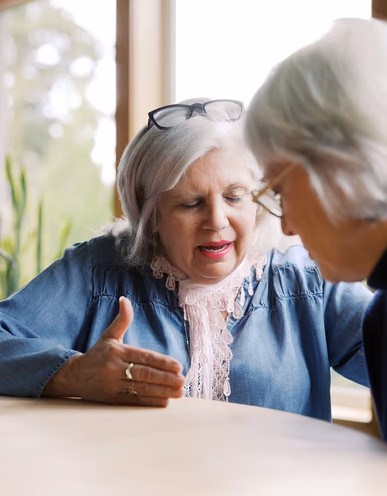 An older woman sitting at a table praying with an older woman.