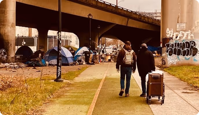 A homeless encampment under a bridge with two men walking on the sidewalk. 