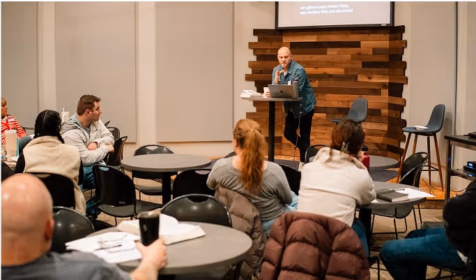 A man stands teaching in front of a classrom of people sitting at tables.