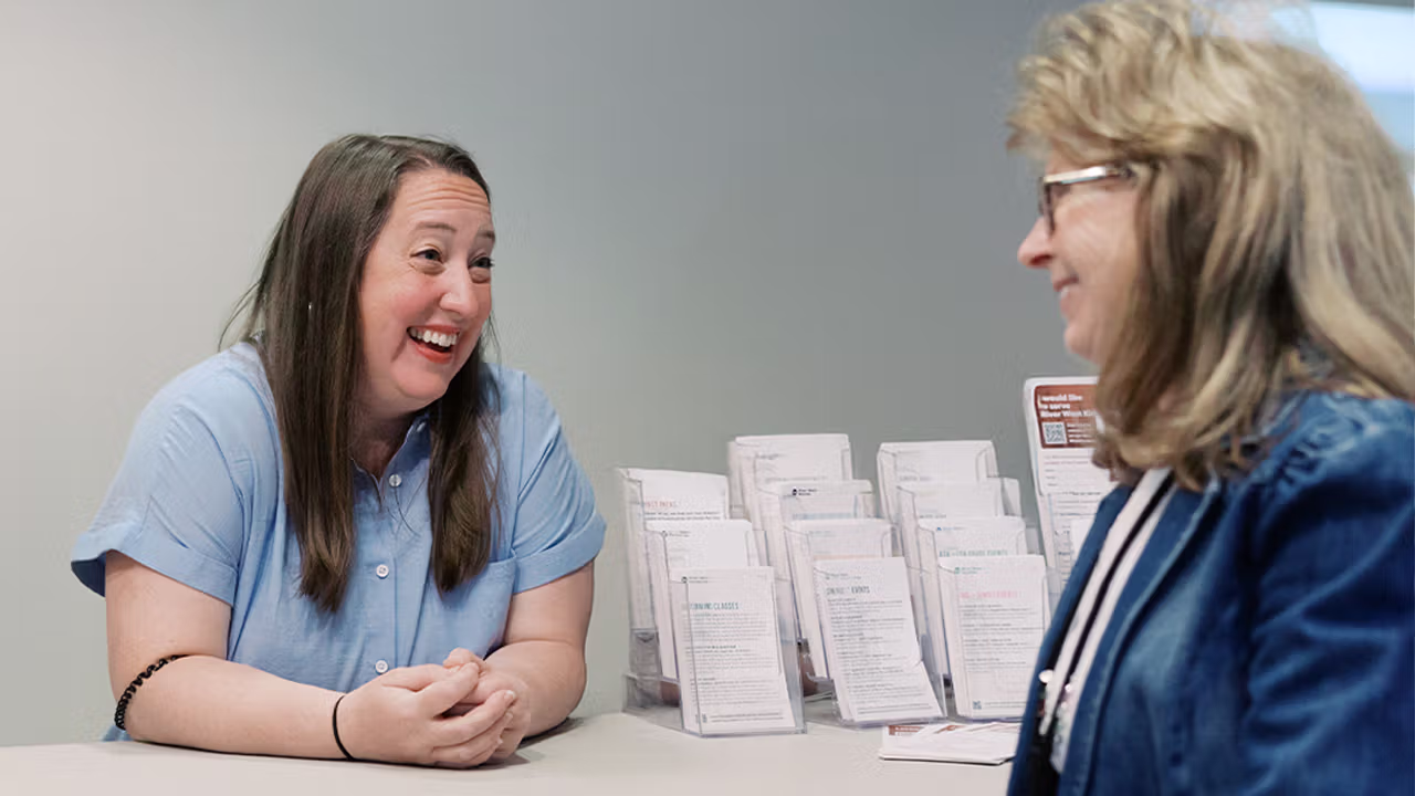 Two women talk at a kiosk next to info pamphlets.