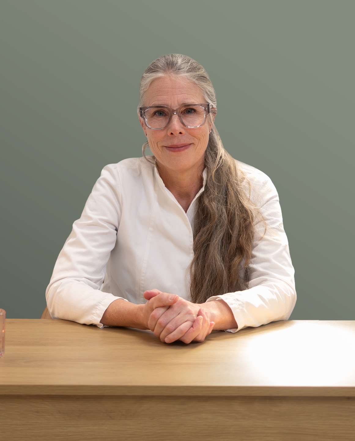 A middle-aged woman named Stina with long gray hair, wearing clear-framed glasses and a white button-up shirt, sitting with her hands clasped and resting on a light wood surface, looking calmly at the camera.