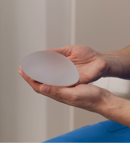 A pair of hands gently holding a smooth, oval-shaped silicone breast implant, showcasing its size, shape, and semi-translucent appearance.