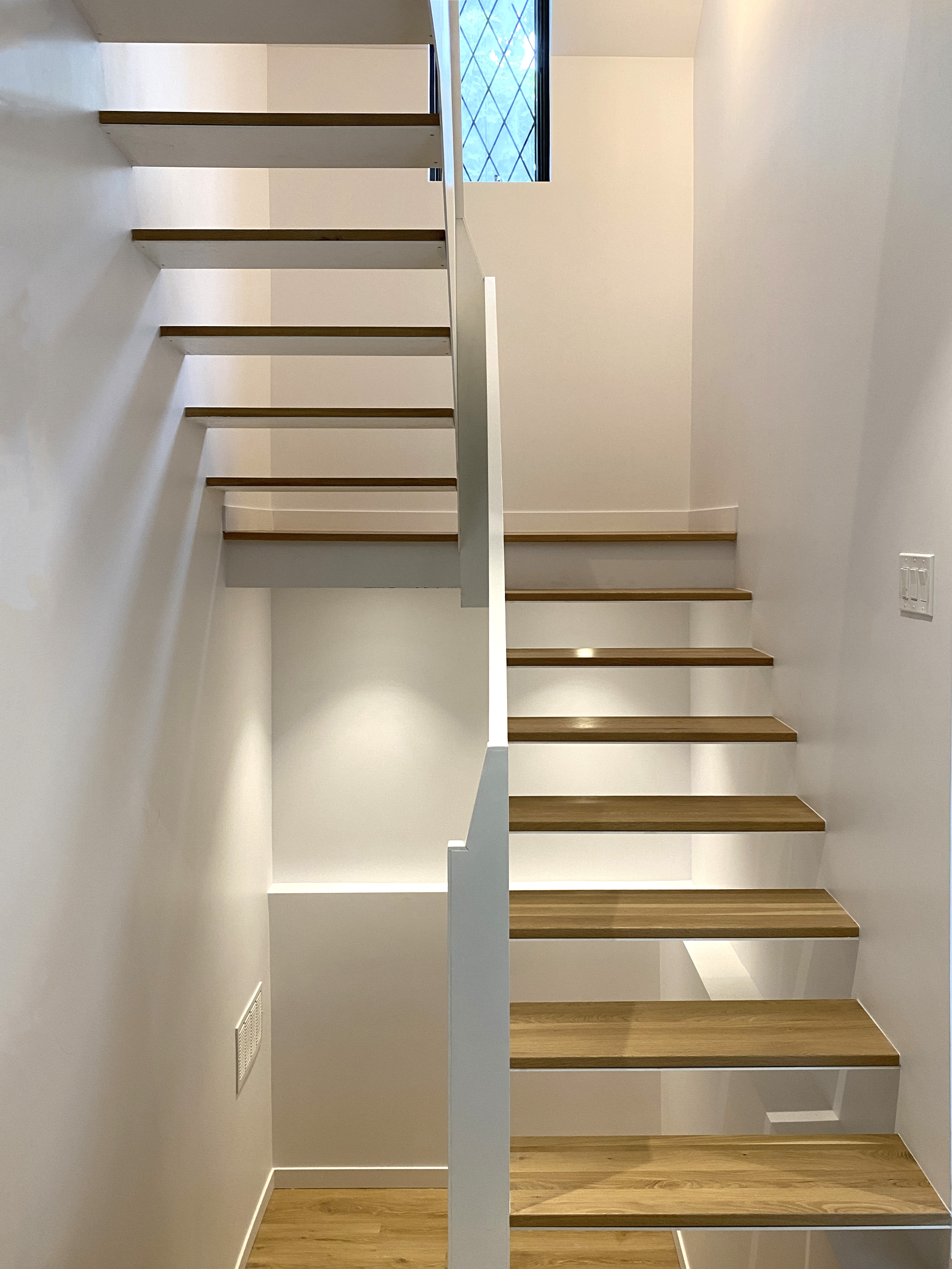 Modern staircase with floating wooden steps and white walls illuminated by natural light from a small window.