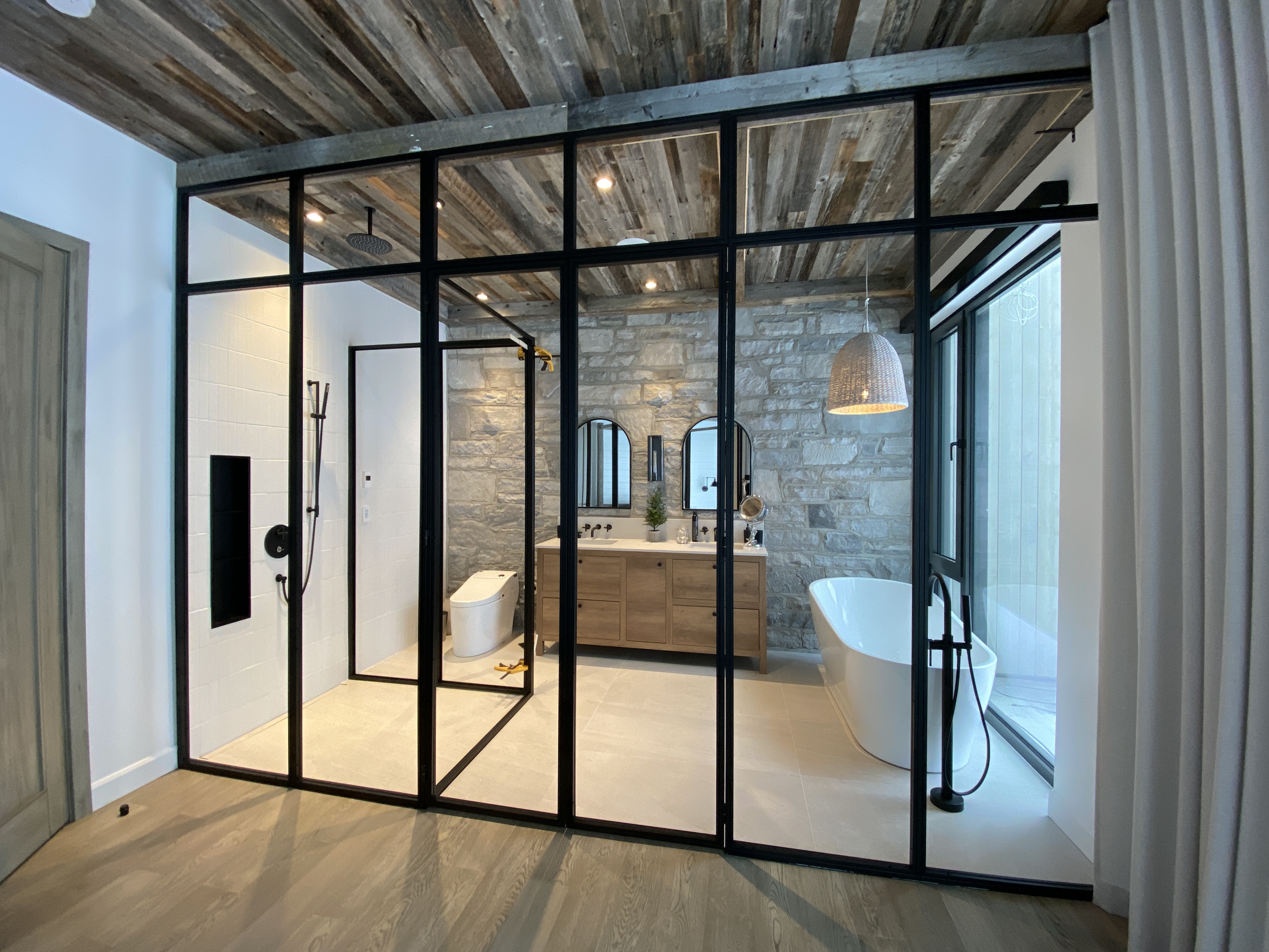 Modern bathroom with glass partition, freestanding white bathtub, wooden vanity with two arched mirrors, stone wall, and wooden ceiling.