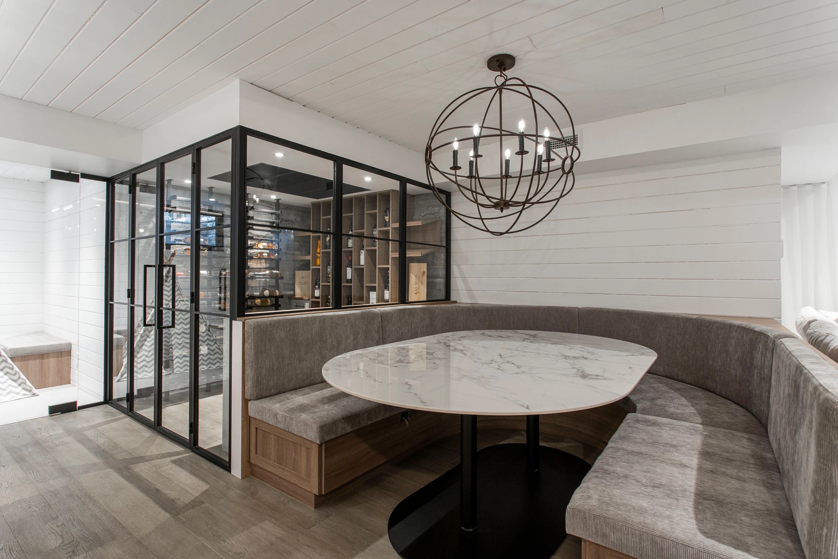 Modern dining nook with gray upholstered curved bench seating, a marble table, and a spherical metal chandelier, next to a glass-enclosed wine cellar.