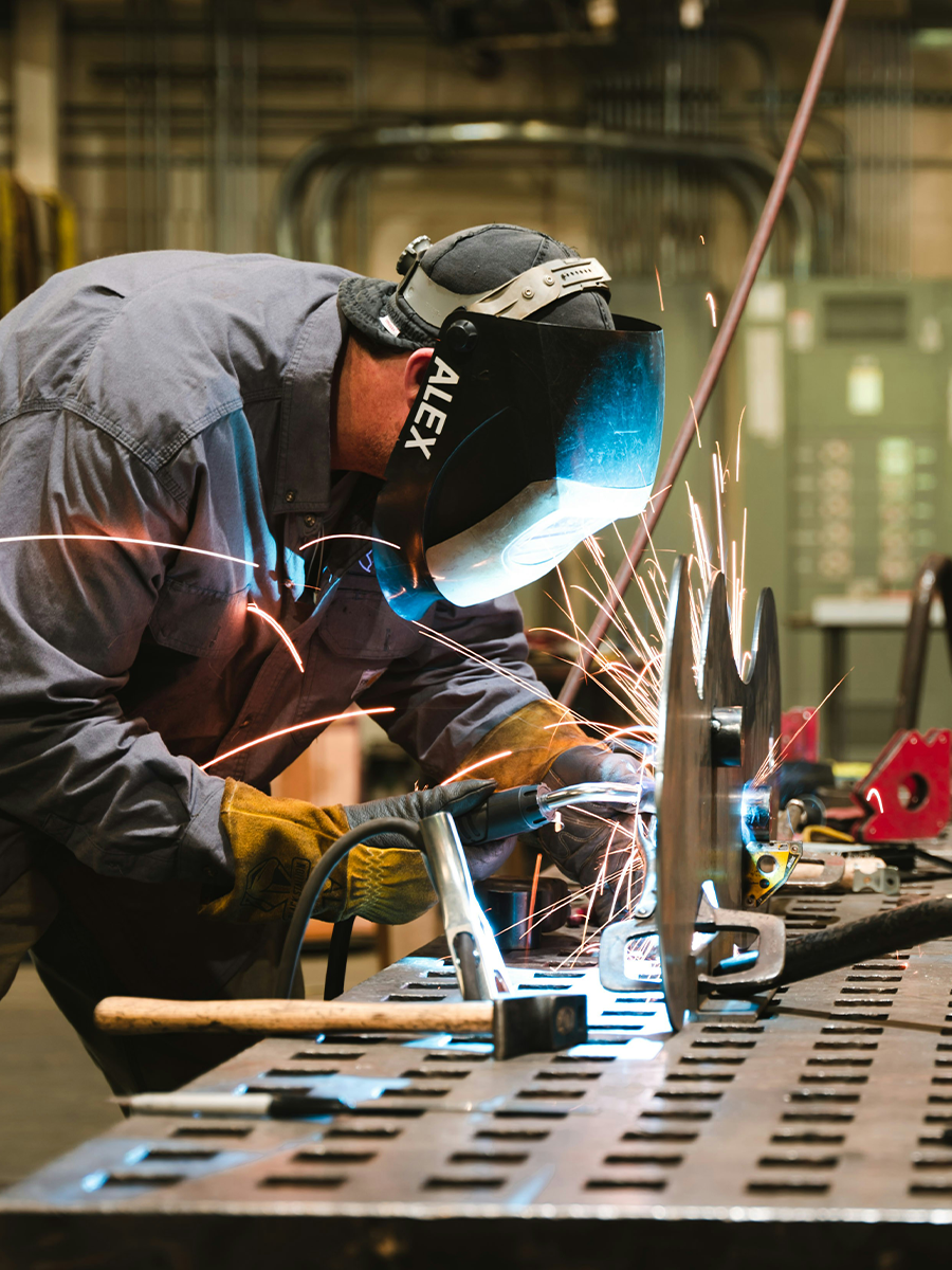 Worker wearing a protective welding mask and gloves, welding metal with sparks flying in an industrial workshop.