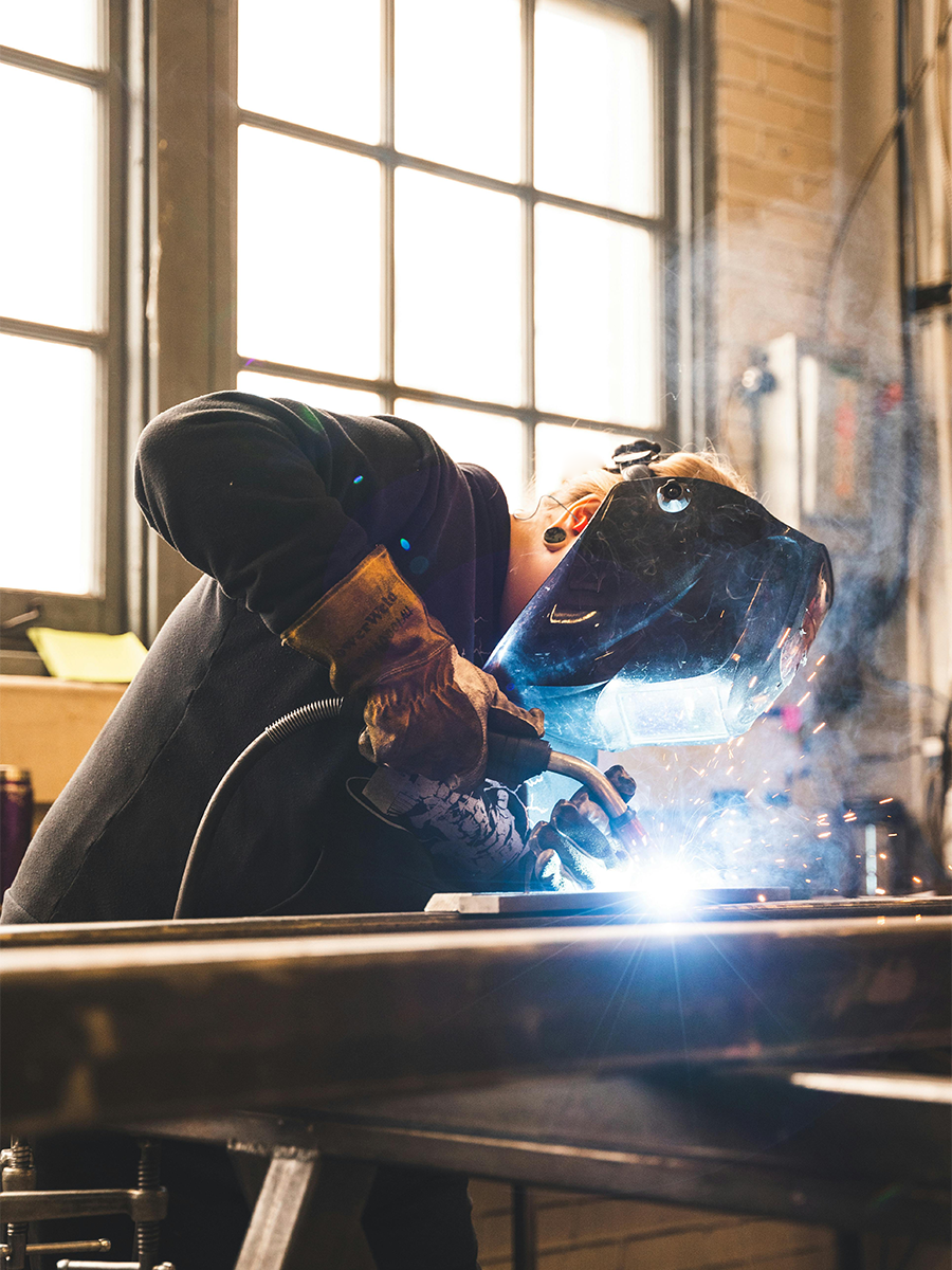 Person wearing protective gloves and welding helmet welding metal in a workshop with sparks and smoke.