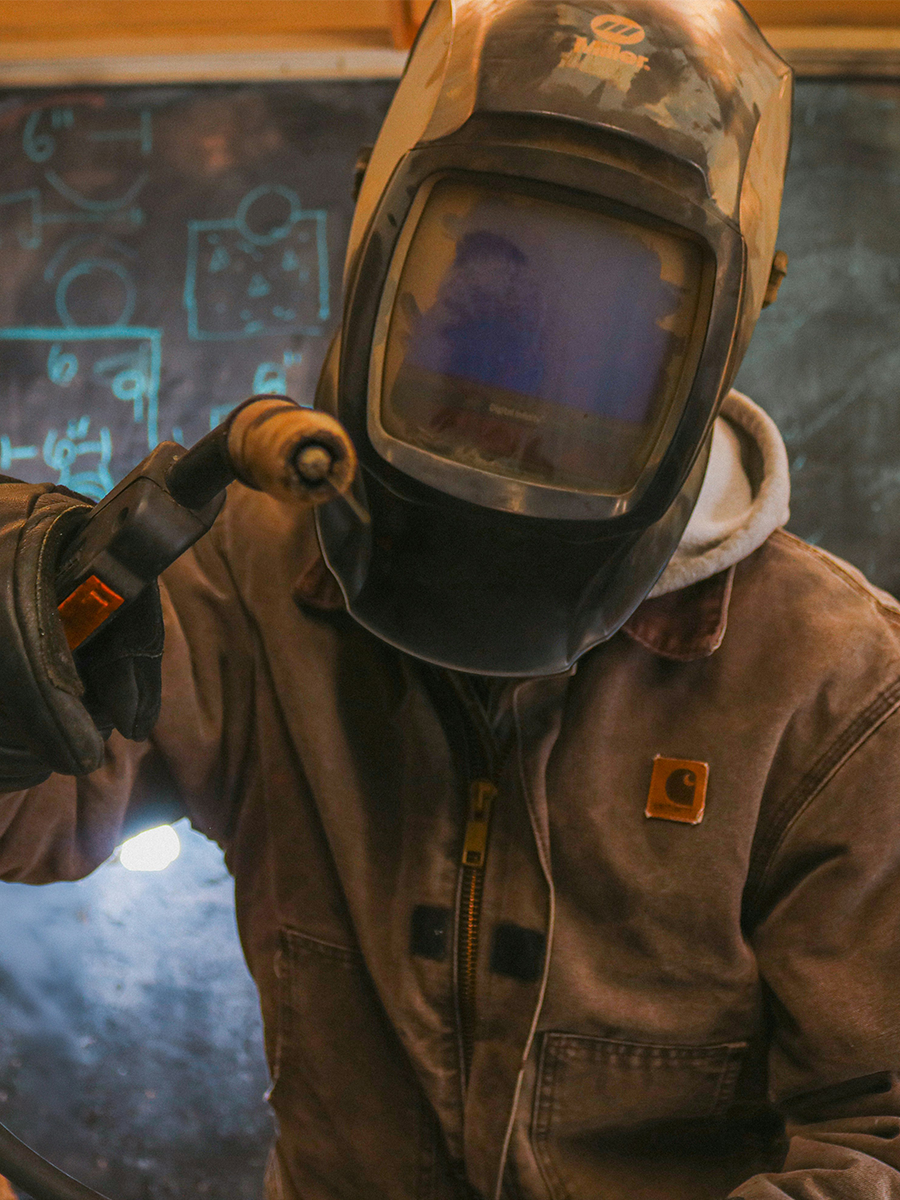 Person wearing a welding helmet and brown jacket holding a welding torch in front of a chalkboard.