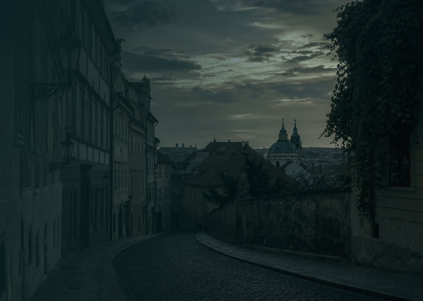 Curved cobblestone street with old buildings and church domes under cloudy evening sky.
