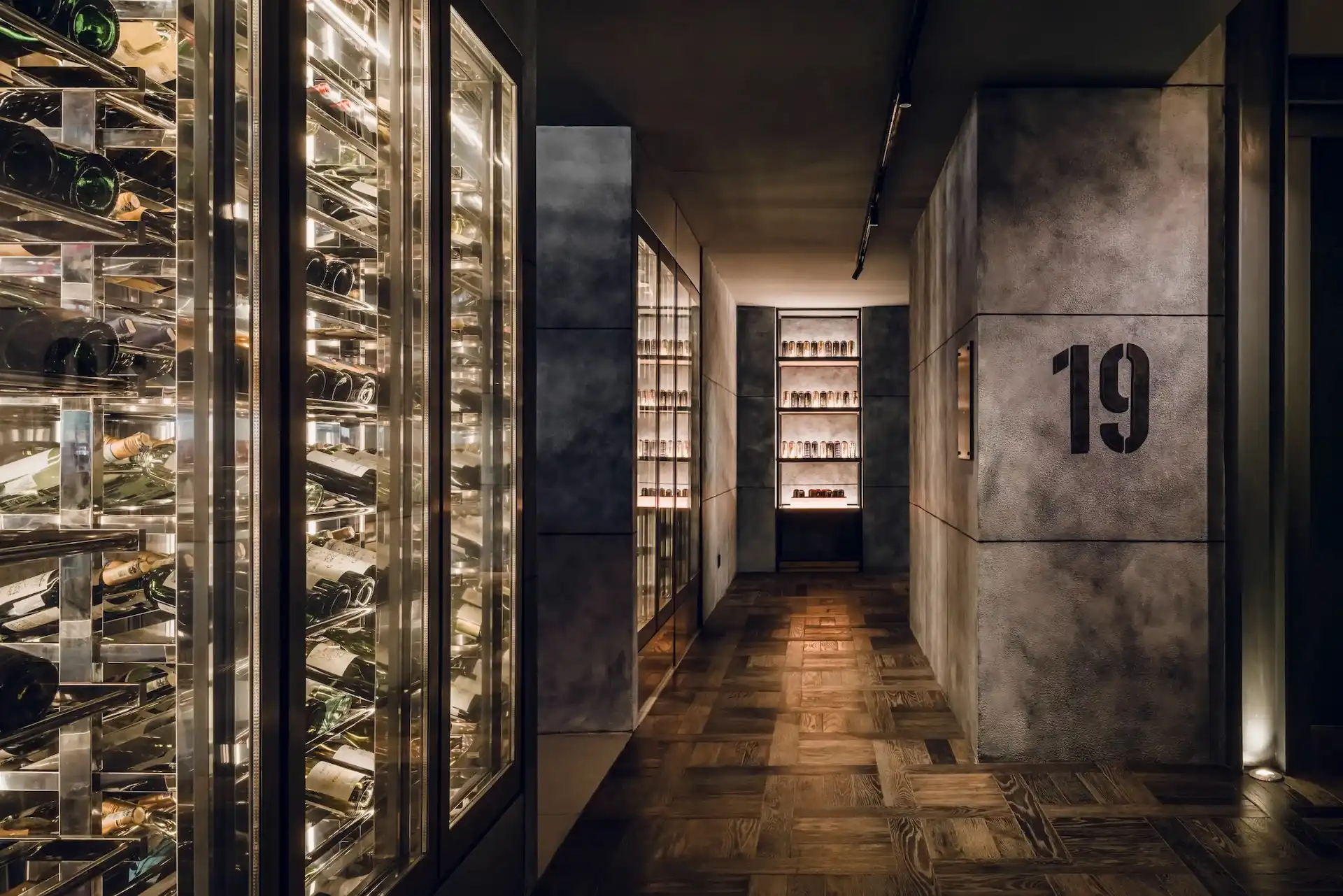 Level 19 lift lobby at Hyatt Centric Kuala Lumpur featuring a glass wine display wall, concrete finish walls, timber parquet flooring and backlit shelving within a contemporary industrial interior.