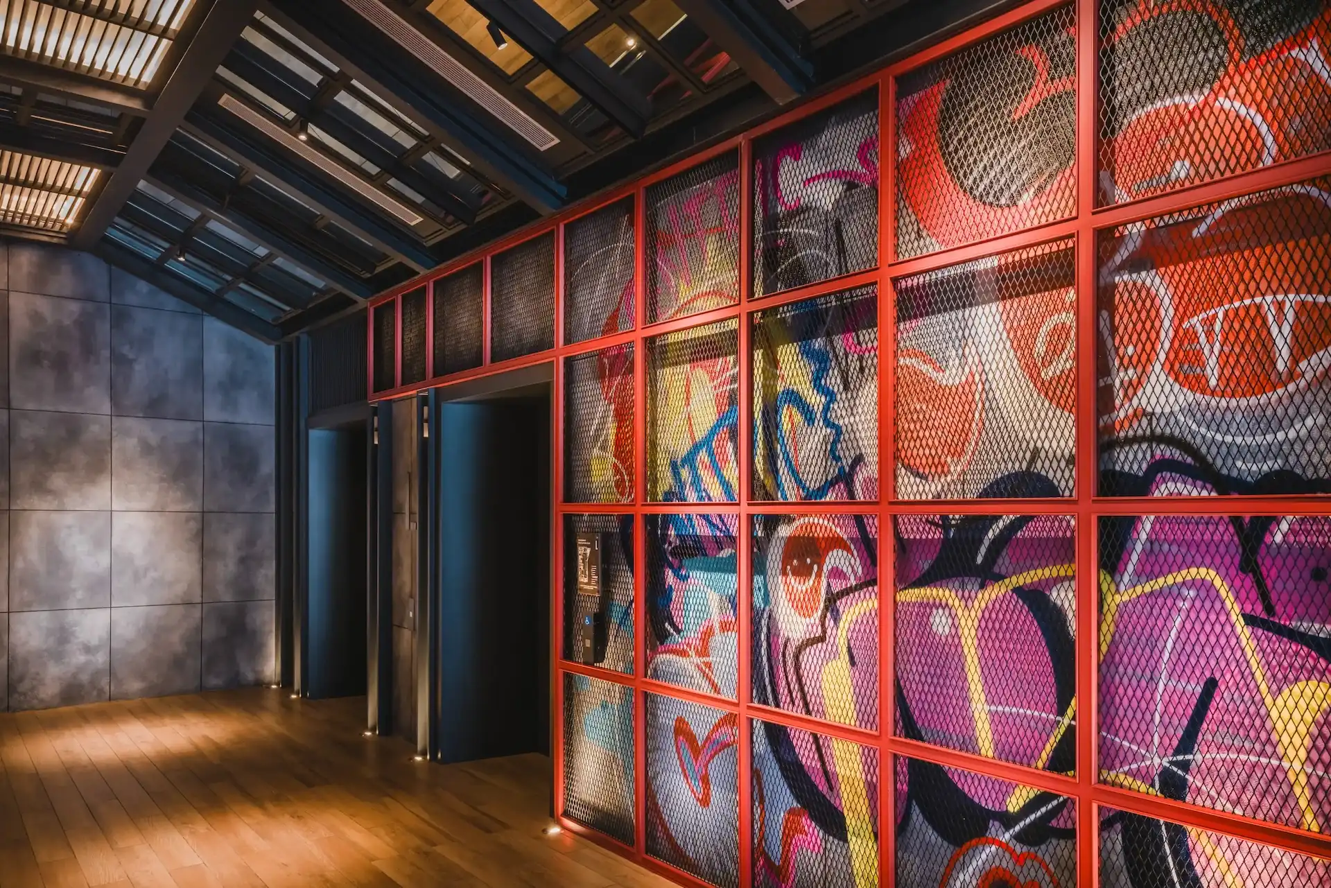 Lift lobby at Hyatt Centric Kuala Lumpur featuring a red steel grid frame, mesh screen panels, graffiti mural backdrop, exposed industrial ceiling and timber flooring.