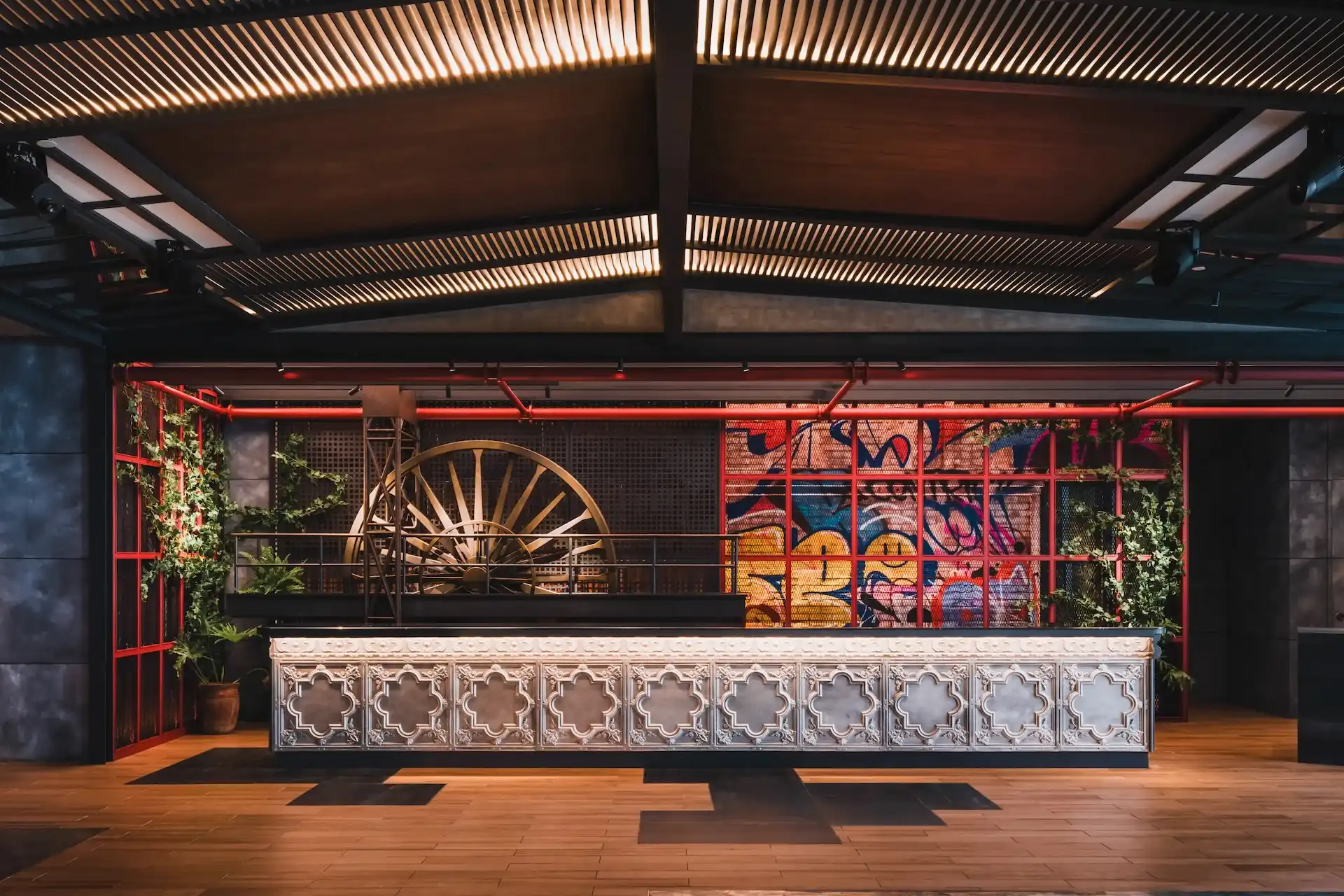 Main reception at Hyatt Centric Kuala Lumpur featuring an ornate reception counter, exposed steel ceiling structure, red metal framework, industrial wheel feature and graffiti mural backdrop inspired by local heritage.