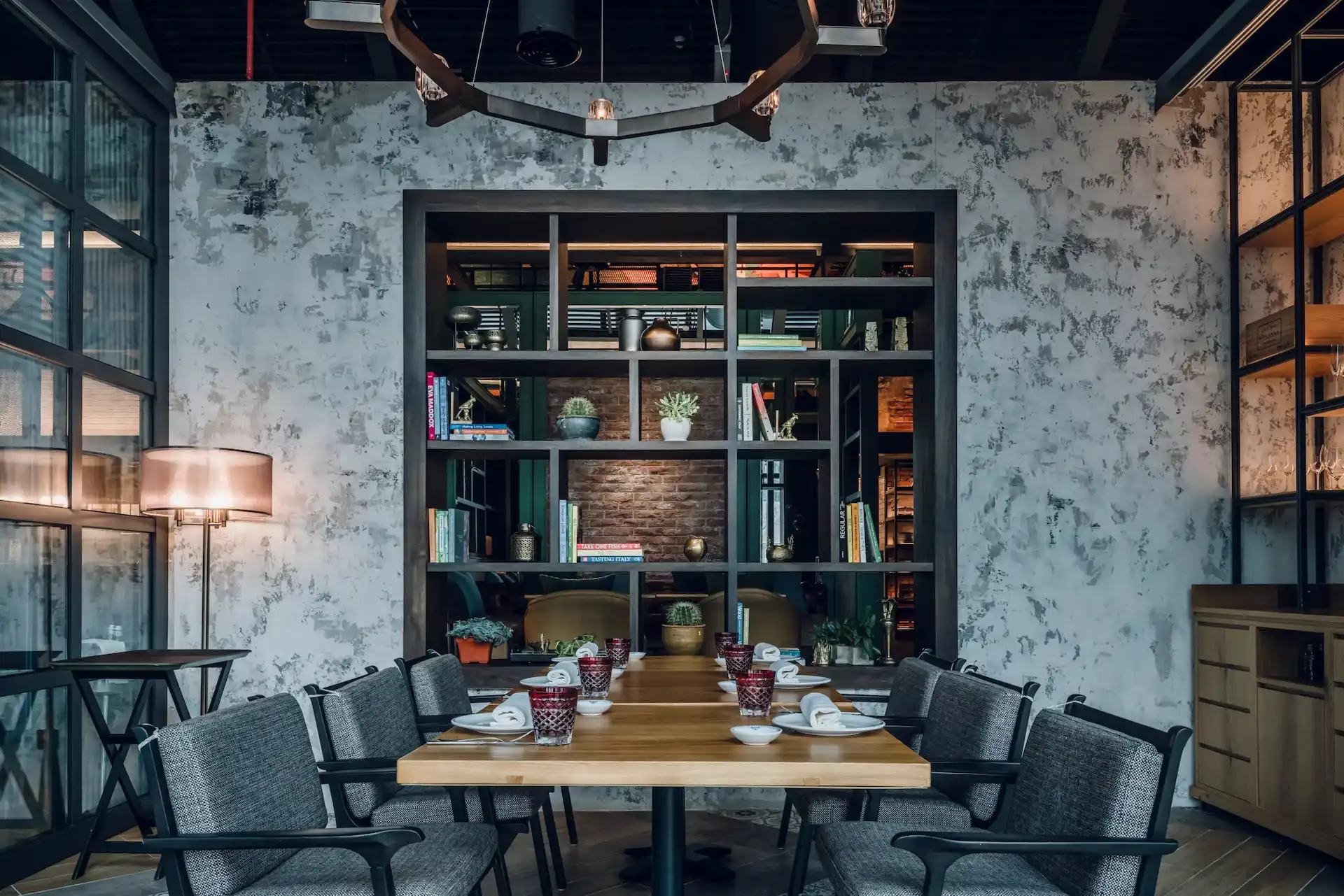Communal dining table in The Gallery at Hyatt Centric Kuala Lumpur with timber tabletop, upholstered armchairs, textured plaster wall and open shelving partition with books, plants and decorative objects.