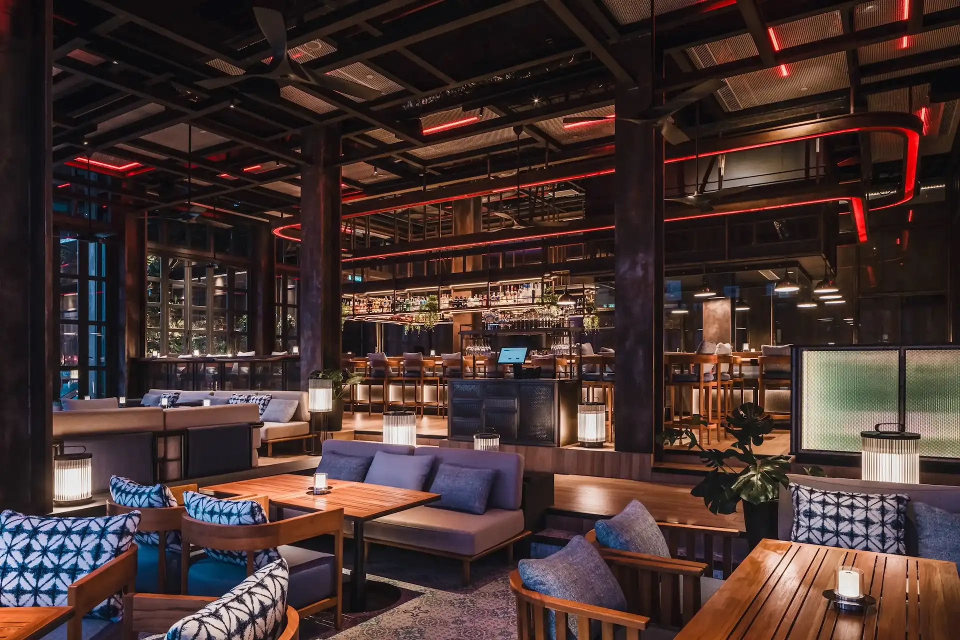 Main lounge area at Level 21 Rooftop Bar in Hyatt Centric Kuala Lumpur featuring timber tables, upholstered sofas, patterned cushions, lantern style table lamps, exposed steel structure with red linear lighting and central bar counter in the background.