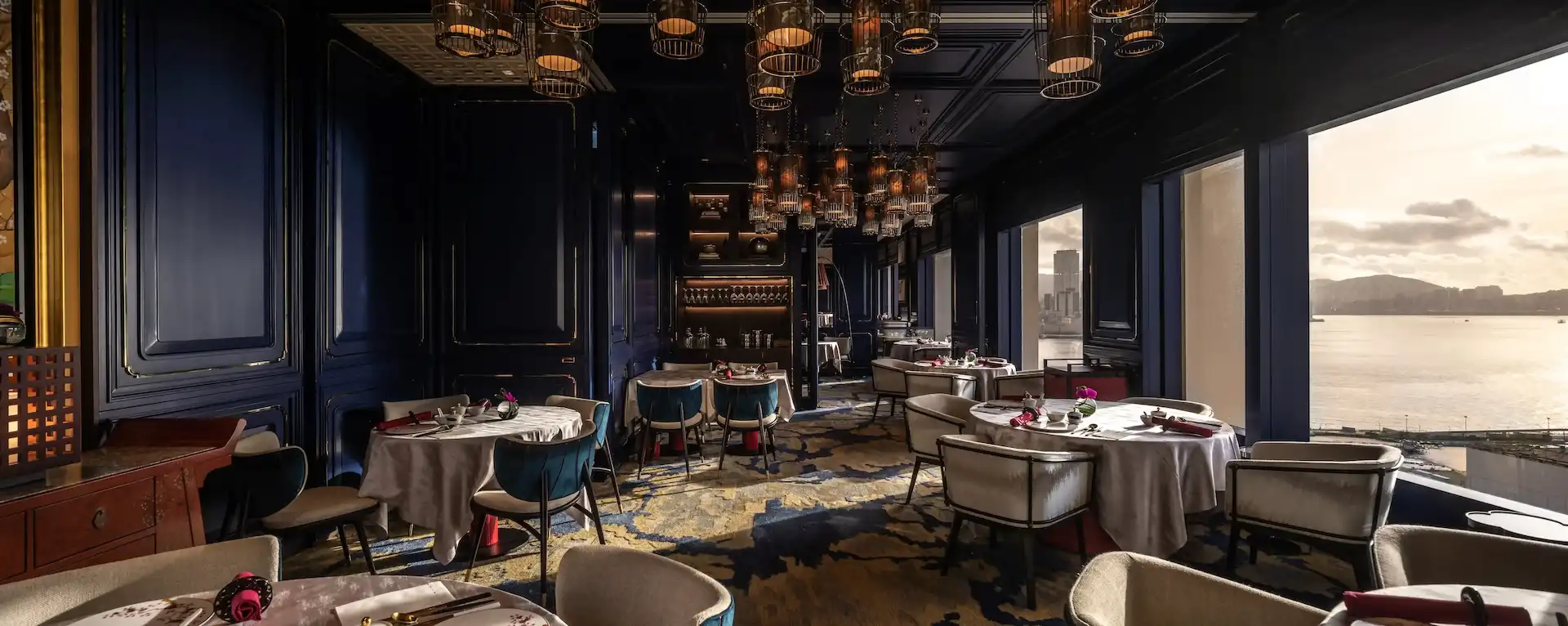 Dining room at Man Wah Mandarin Oriental Hong Kong featuring navy panelled walls with gold detailing, lantern chandeliers, round tables with white tablecloths and red napkins, patterned blue and gold carpet, and panoramic harbour views through full height windows.