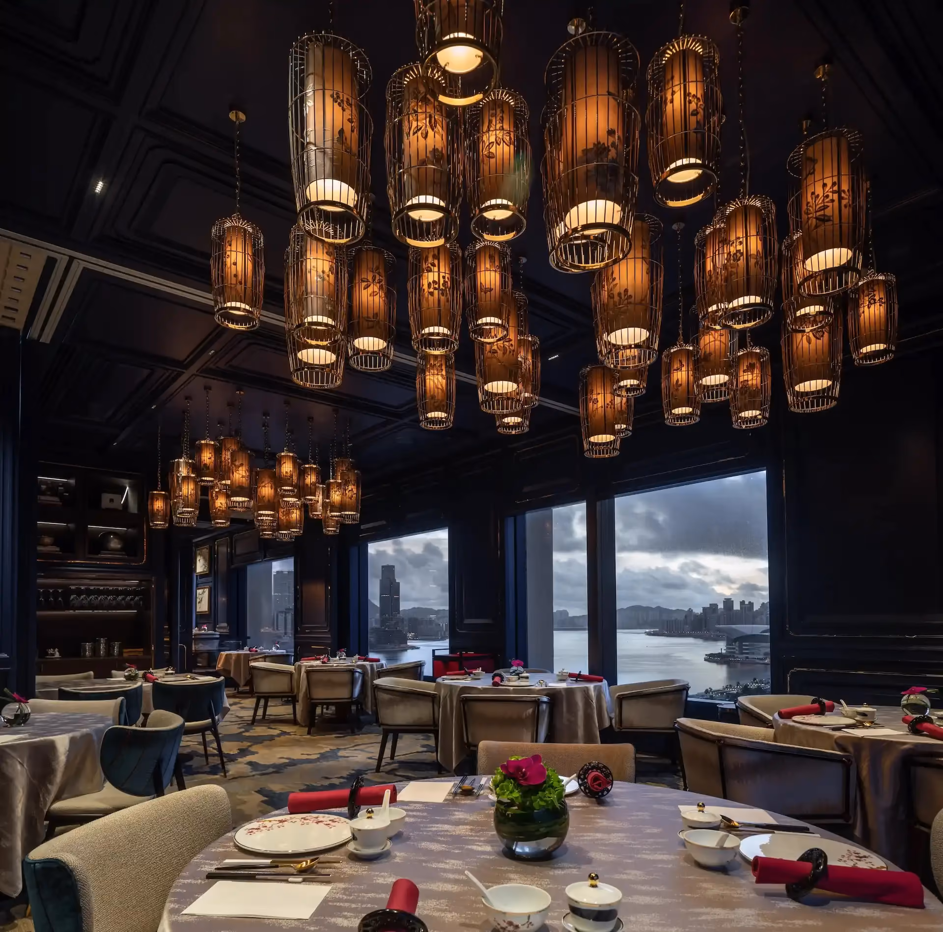 Dining room at Man Wah Mandarin Oriental Hong Kong featuring suspended lantern chandeliers, navy panelled walls, round tables with white tablecloths and red napkins, and panoramic views over Victoria Harbour through full height windows.