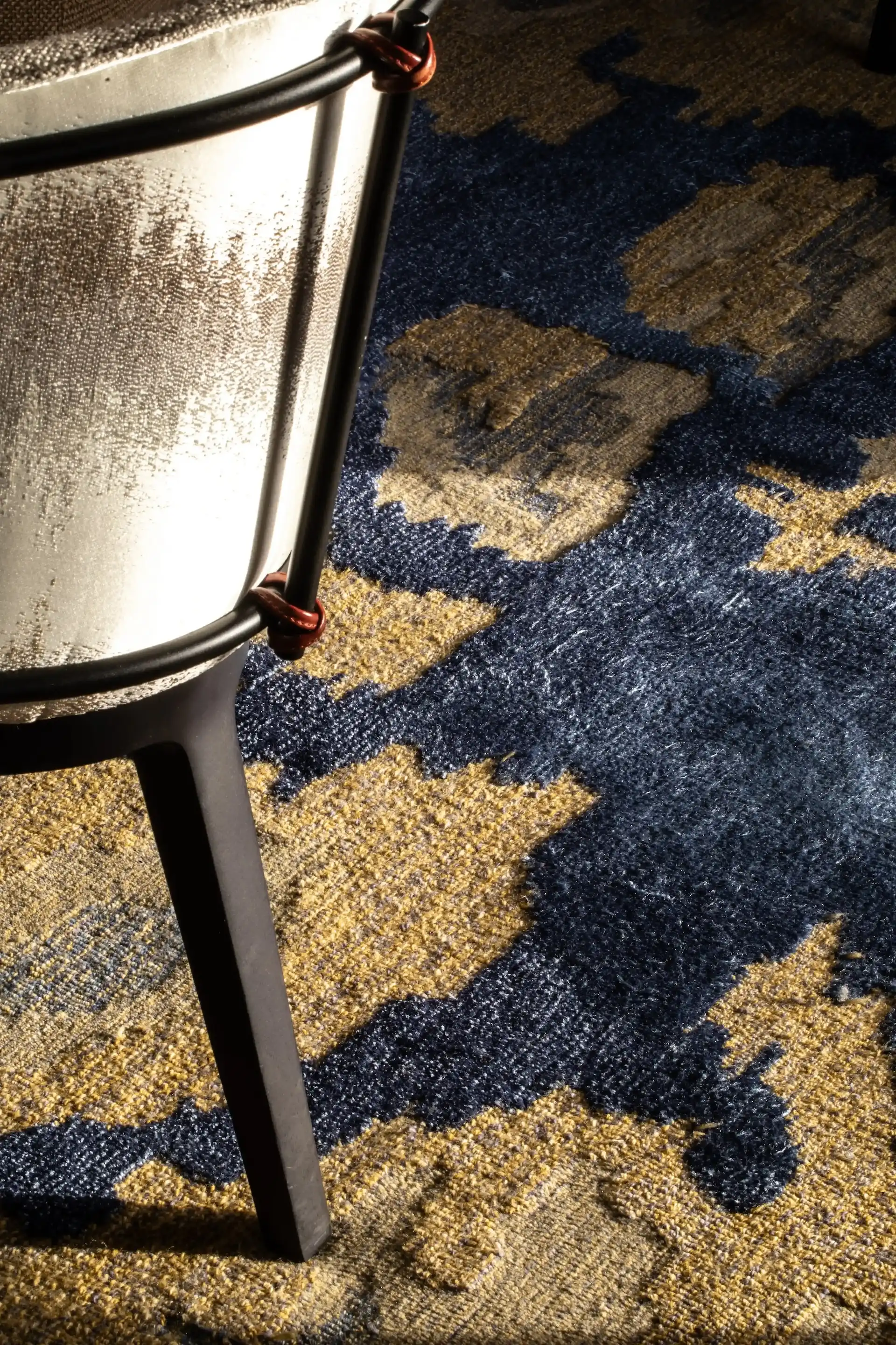 Close up detail at Man Wah Mandarin Oriental Hong Kong showing upholstered dining chair with metal frame and leather ties positioned on blue and gold patterned carpet with abstract floral motif.