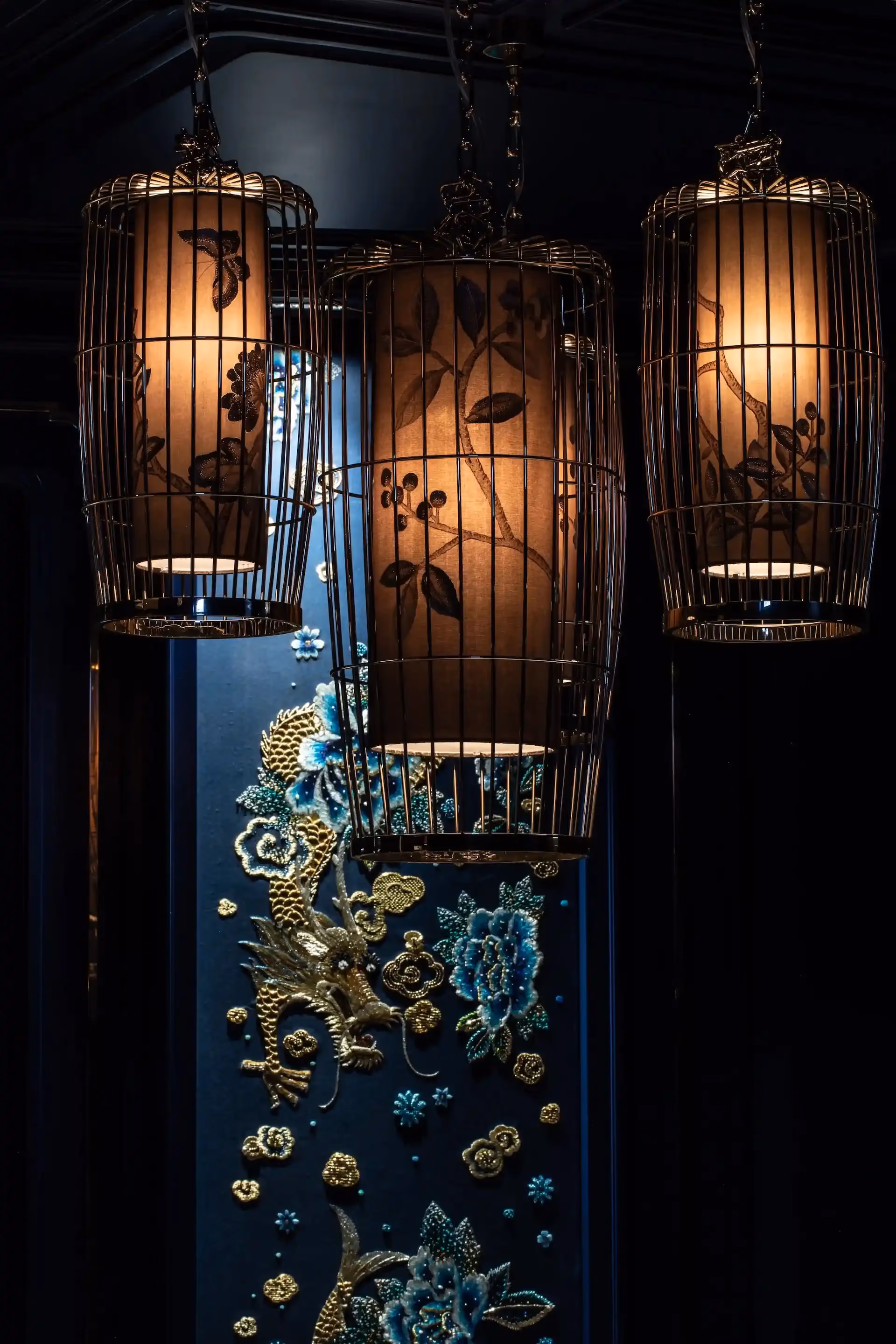 Close up detail at Man Wah Mandarin Oriental Hong Kong showing suspended lantern chandeliers with warm glow, metal cage frames and floral motifs, set against navy wall panel with embroidered dragon and floral artwork.
