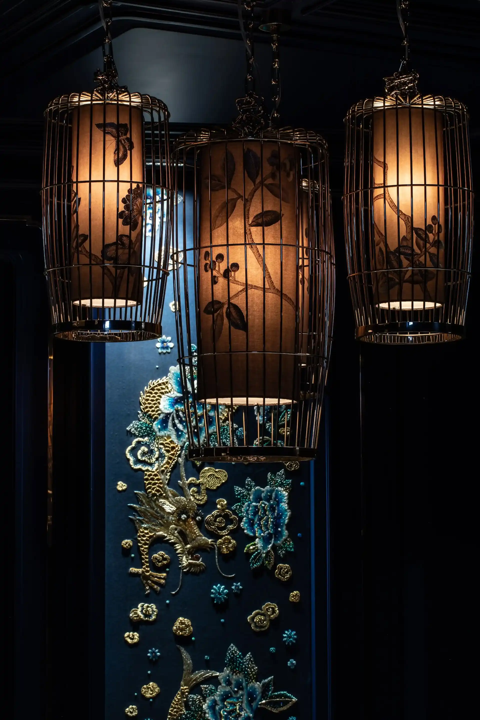 Close up detail at Man Wah Mandarin Oriental Hong Kong showing suspended lantern chandeliers with warm glow, metal cage frames and floral motifs, set against navy wall panel with embroidered dragon and floral artwork.
