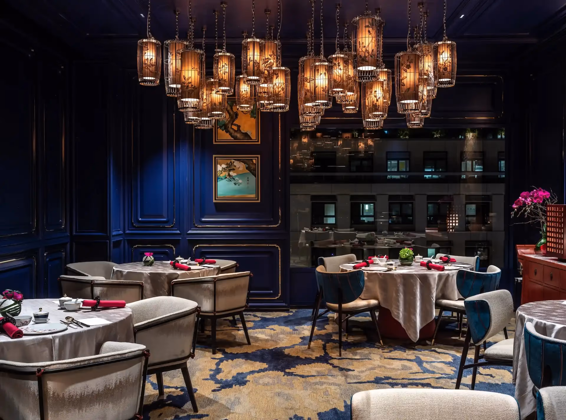 Dining room at Man Wah Mandarin Oriental Hong Kong featuring clustered lantern chandeliers with warm illumination, navy blue panelled walls with gold trim, round dining tables with white tablecloths and red napkins, patterned carpet in blue and gold, and framed artwork.